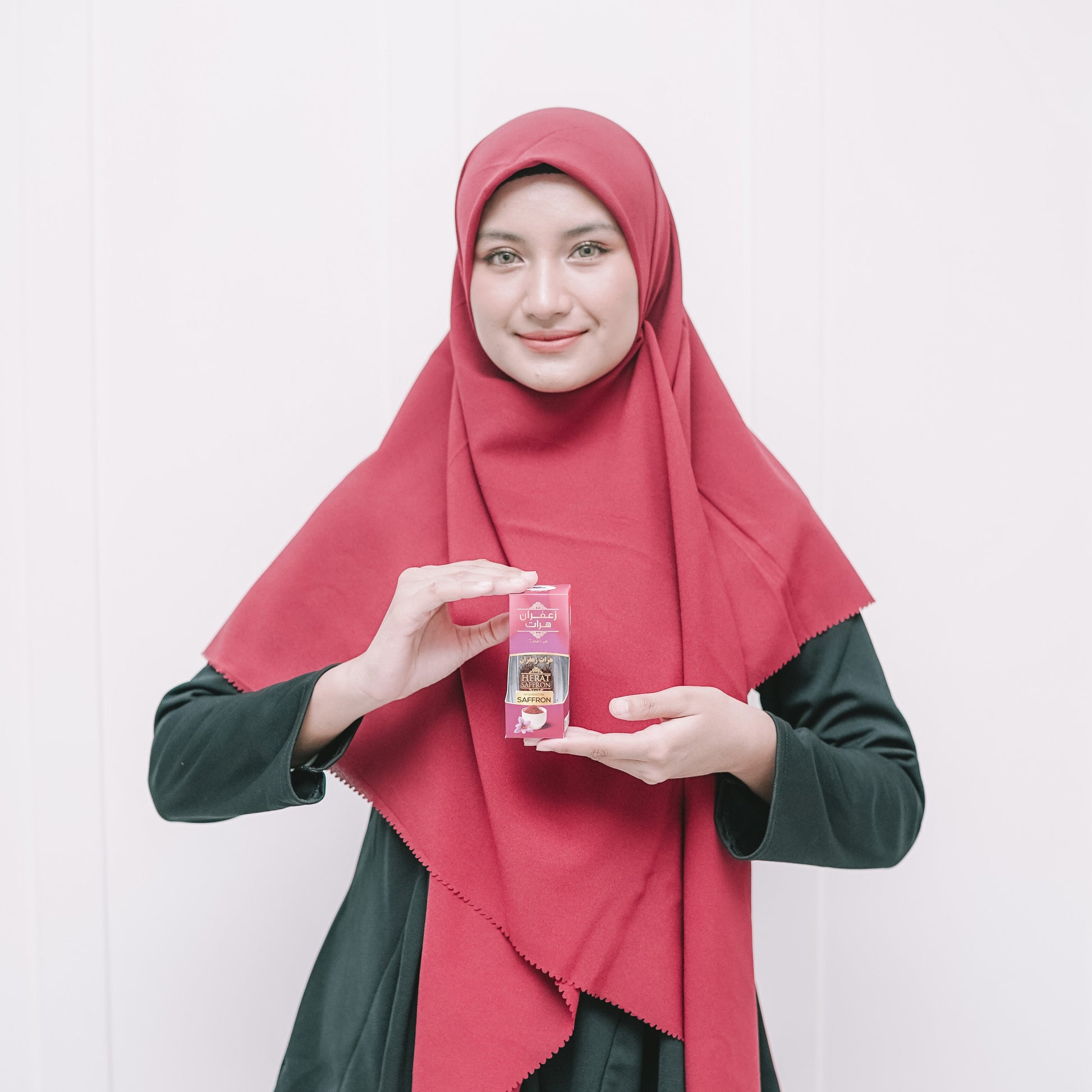 Woman wearing a red hijab and green outfit holding a product against a white background