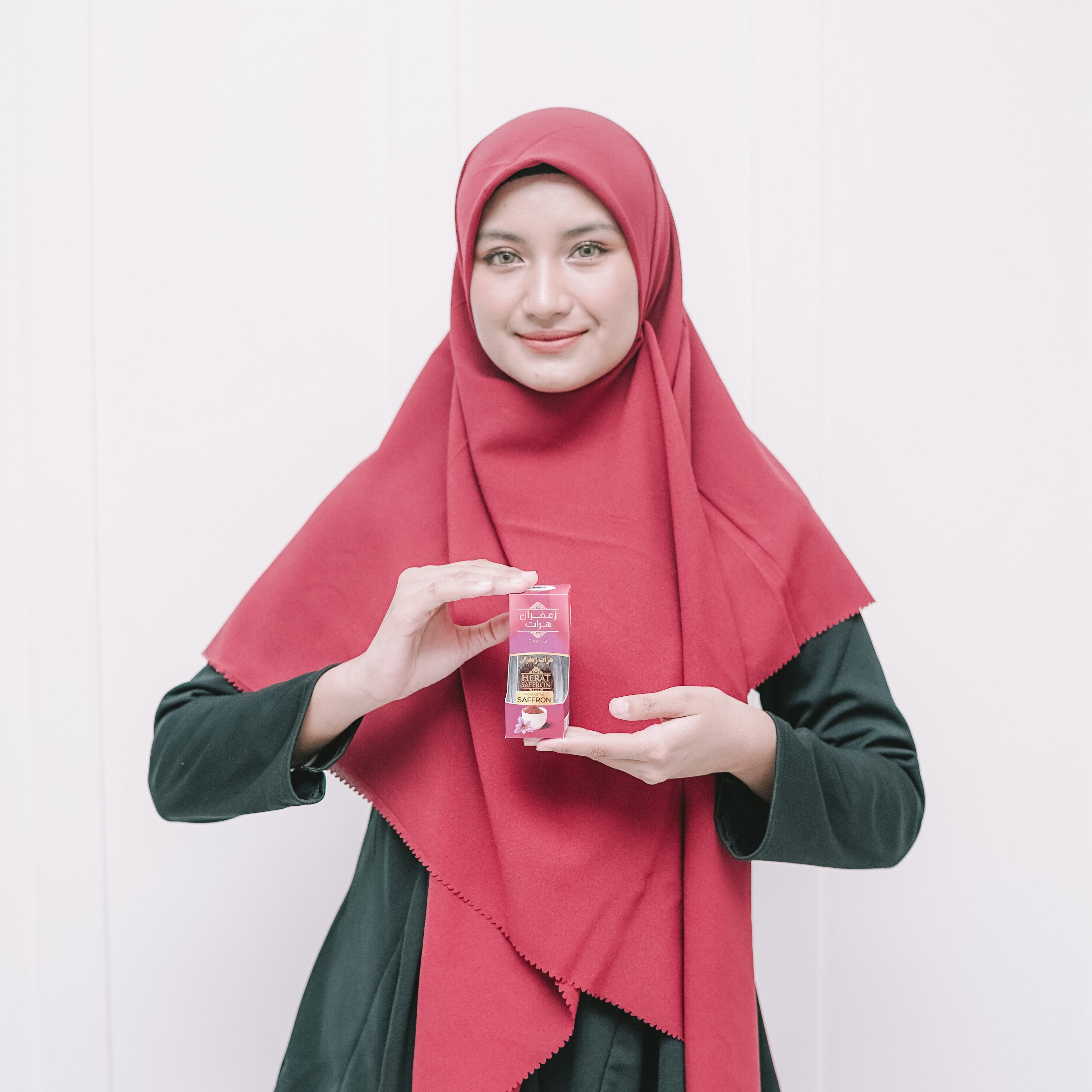 Woman wearing a red hijab and green outfit holding a product against a white background