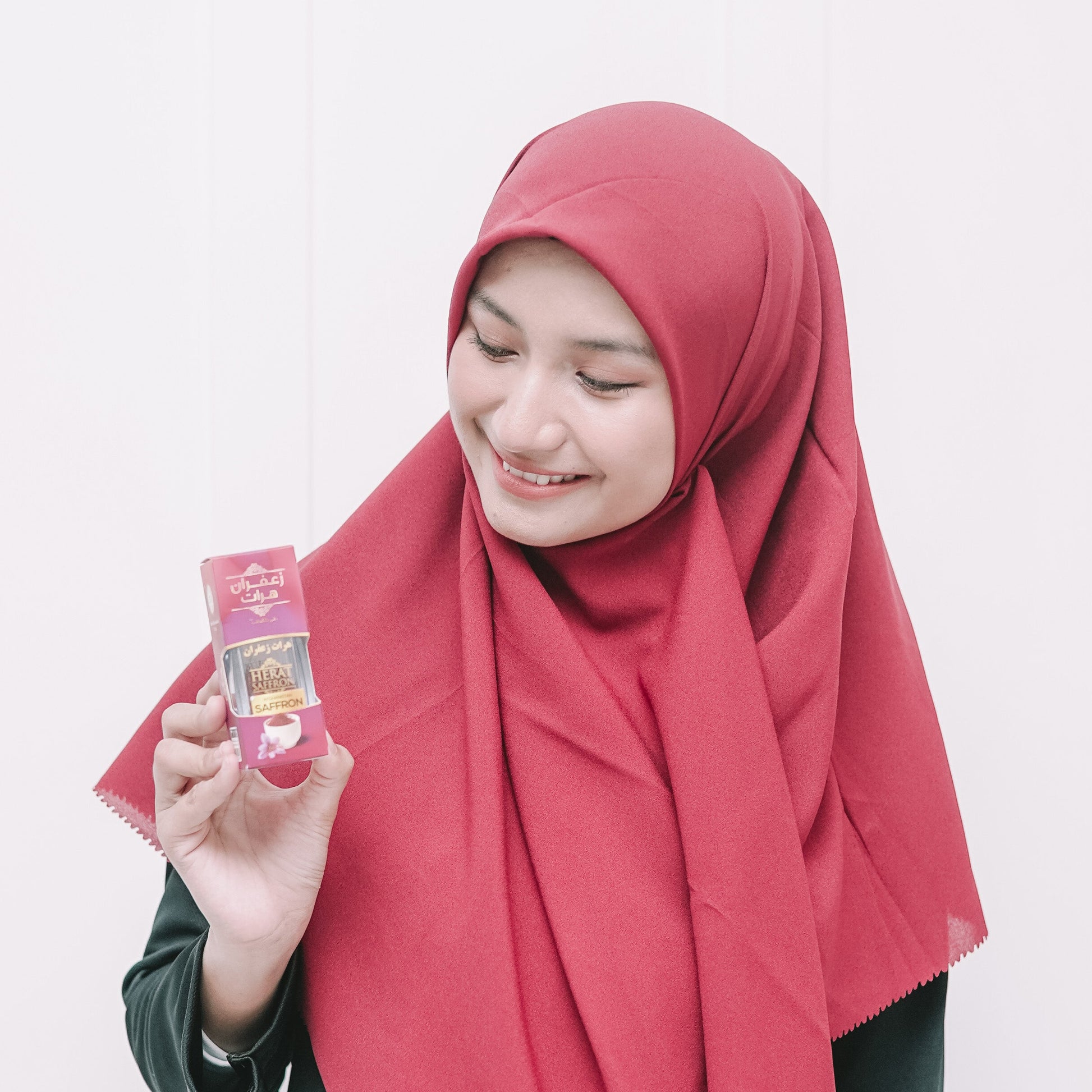 Woman wearing a red hijab and green outfit holding a small package against a white background
