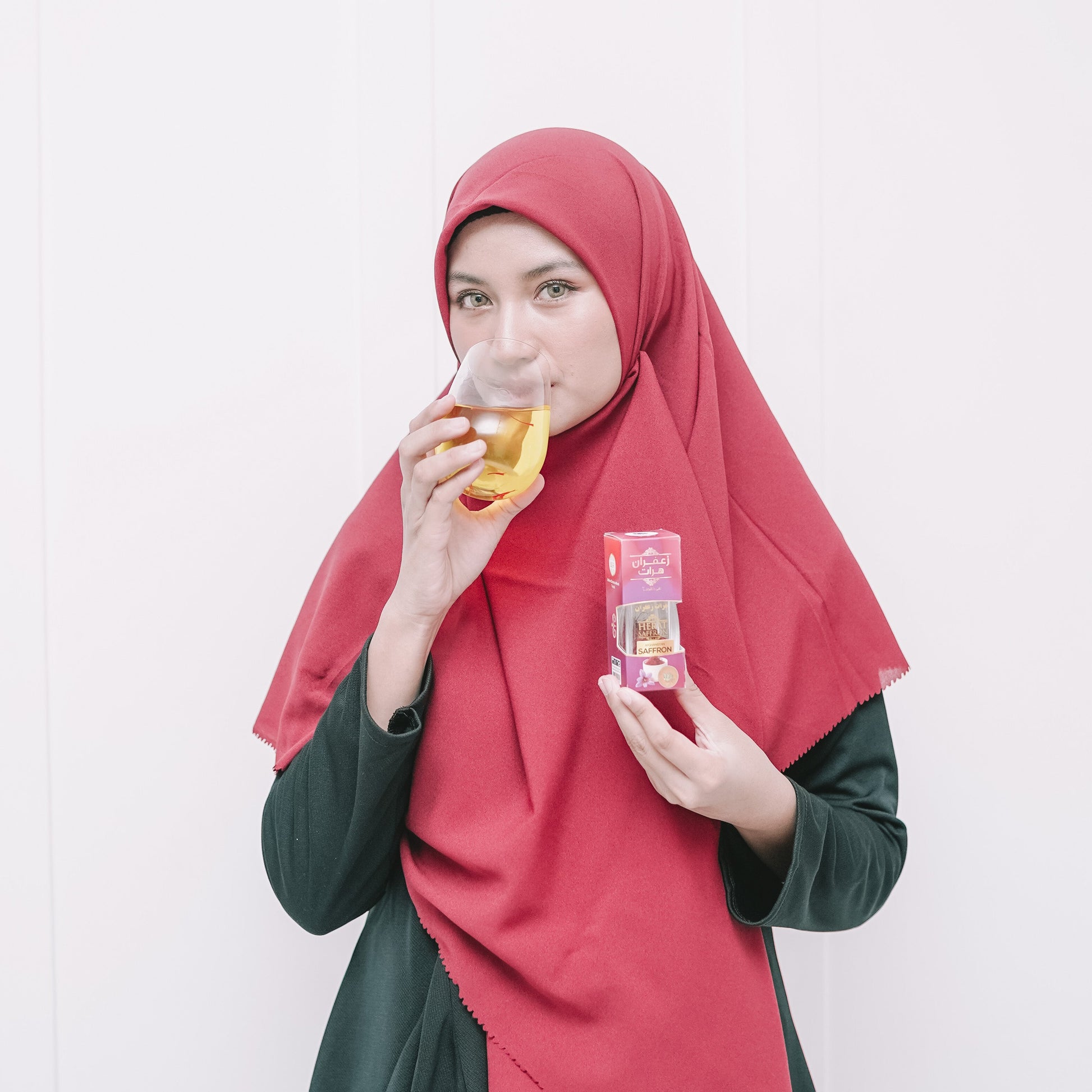 Woman wearing a red hijab and green outfit, holding a drink and a snack against a white background