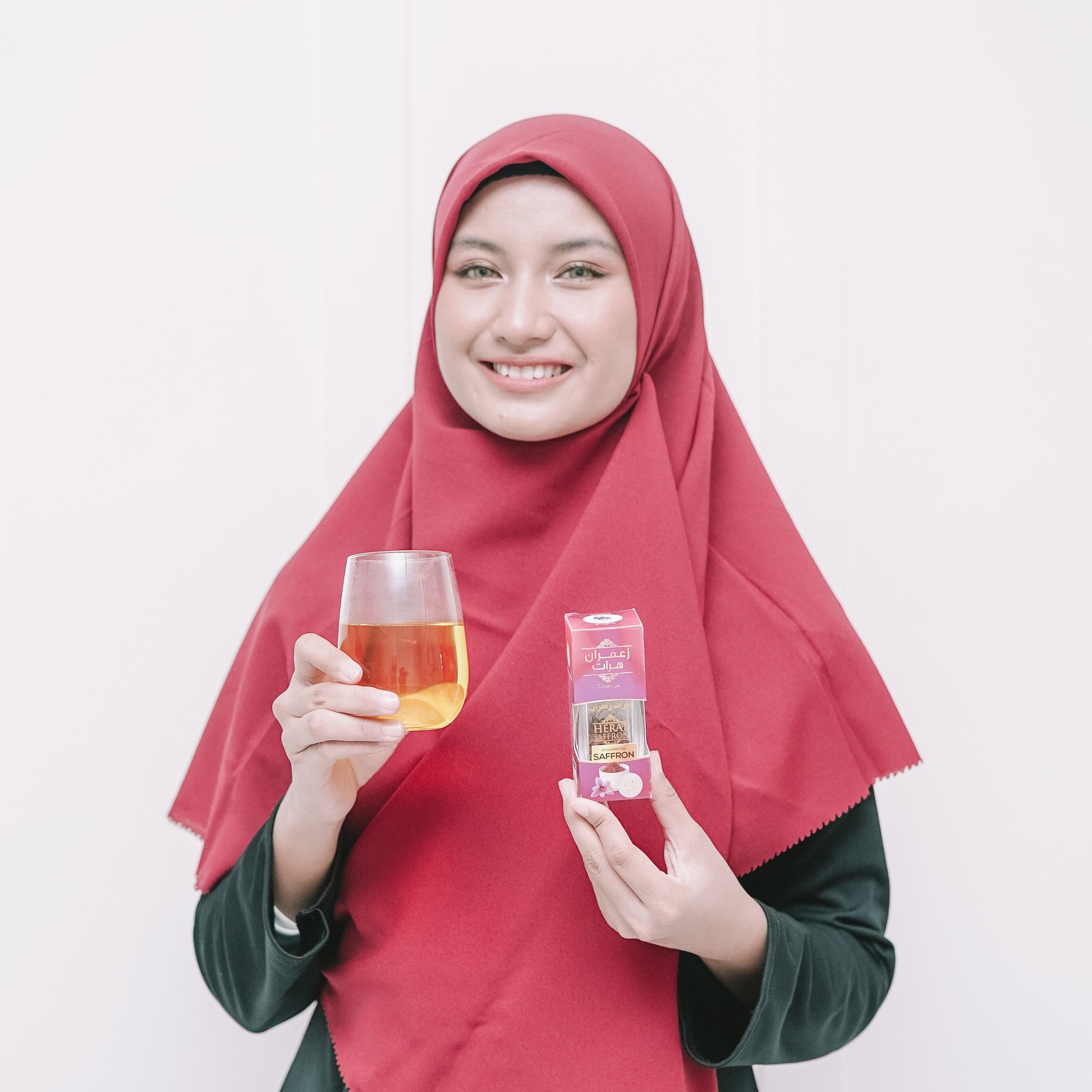 Woman wearing a red hijab holding a glass of tea and a small bottle against a white background
