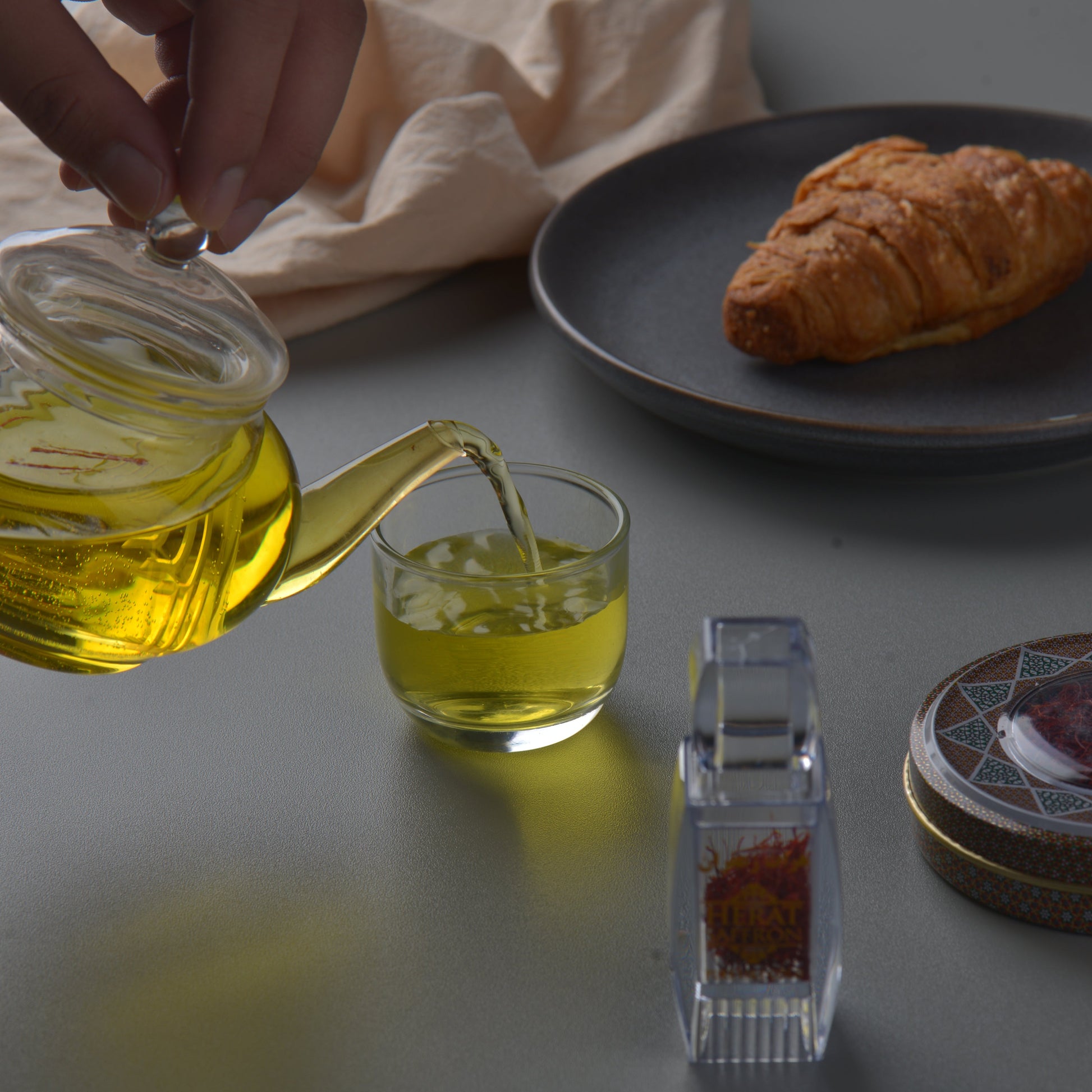 Person pouring oil from a bottle into a glass, with a croissant on a plate in the background.